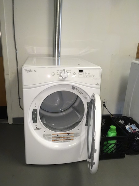 A white dryer sitting on a gray floor with a silver dryer tube running up the wall to the outside of the room. The dryer door is open. There is a black milk crate with a green bottle of Gain laundry soap in it on the floor next to the dryer.