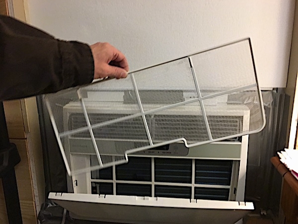 A white Frigidaire window heat pump unit inside of a window with the front panel open and a person pulling out a screen-like filter.