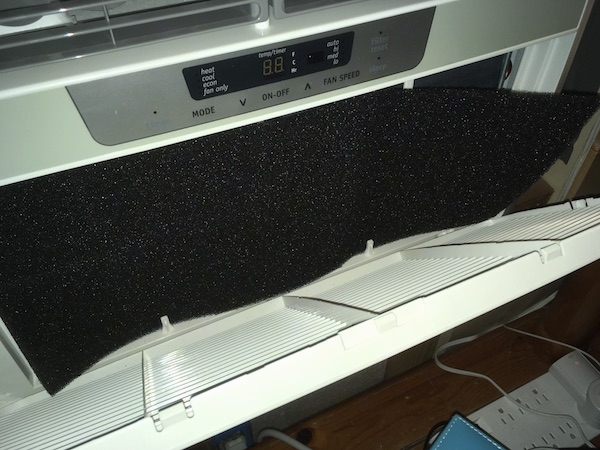 A white Frigidaire window heat pump unit inside of a window with the front panel open showing a gray sponge-like filter.