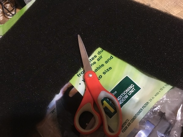 A gray sponge-like foam filter laying on a table on top of the plastic packaging bag with a pair of red scissors next to it.