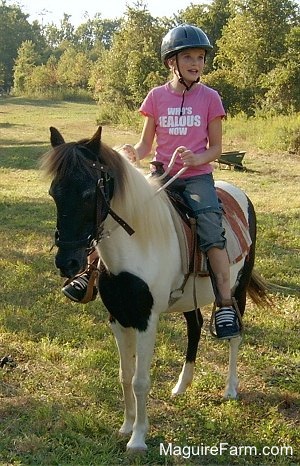 A smiling girl in a pink shirt and blue helmet is sitting on the back of a brown and white paint pony holding the reins out in a feild. There is a wheel barrel in the distance sitting out in the grass.