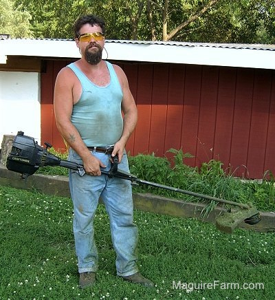 Bob standing in front of the red chicken coop wearing yellow safty glasses holding a weedwacker