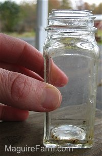 There is a clear little jar on a wooden table outside on a porch. There is a person holding the jar