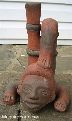 A Clay African Statue is on a stone porch. The Statue is of a person with there head and arms on the ground. Its legs are in the air. One of the feet are touching the head of the african statue