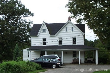 The side view of a white farm house with a wrap around stone porch and a black Toyota Avalon and a light blue Toyota Sienna parked in front of it. There is a golf cart parked on the porch.