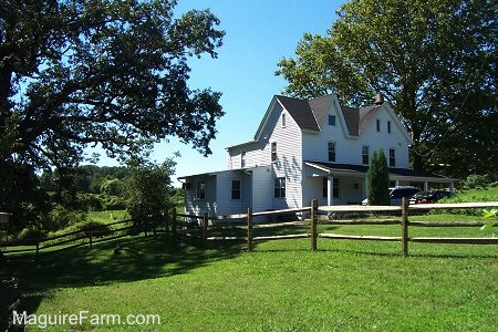 A backside perspective of a white farm house with a wrap around porch. There is green grass and a split rail fence around the house.