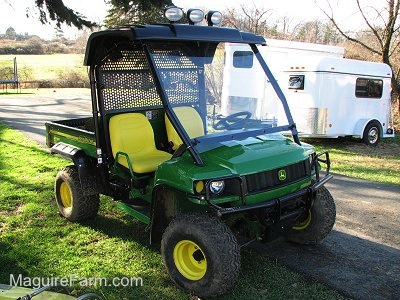A green John Deere HPX 4x4 Gator parked along side of a driveway with two white horse trailers parked on the other side.