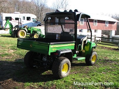 The backside of a John Deere HPX 4x4 Gator with a second John Deere tractor, an RV camper and a red barn in the distance.