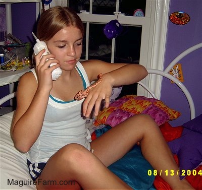 A blonde-haired girl in a white shirt is on the phone while holding an orange and white snake. The walls in the room are purple and she is sitting on her white bed.