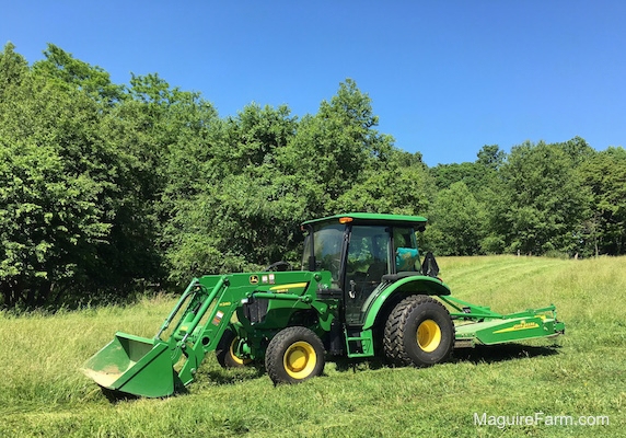 John Deere 5065E Compact Tractor and Front End Loader with a cutting deck out in a green grassy fields with trees behind it